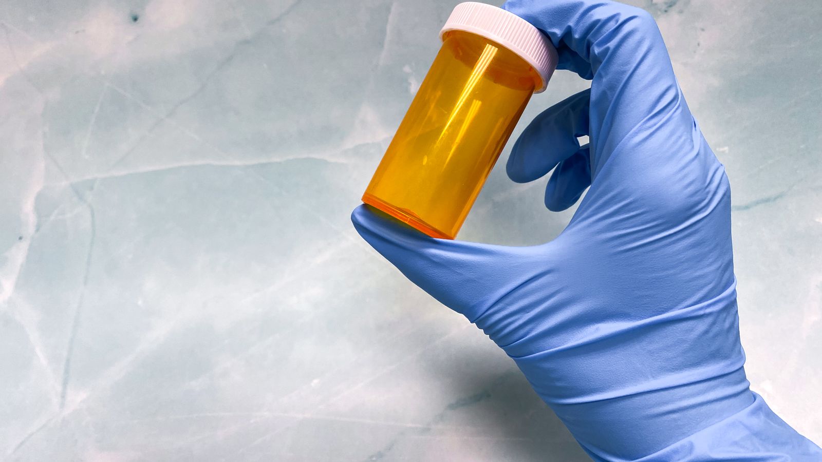 Close-up of a gloved hand holding an empty yellow prescription vial against a gray background
