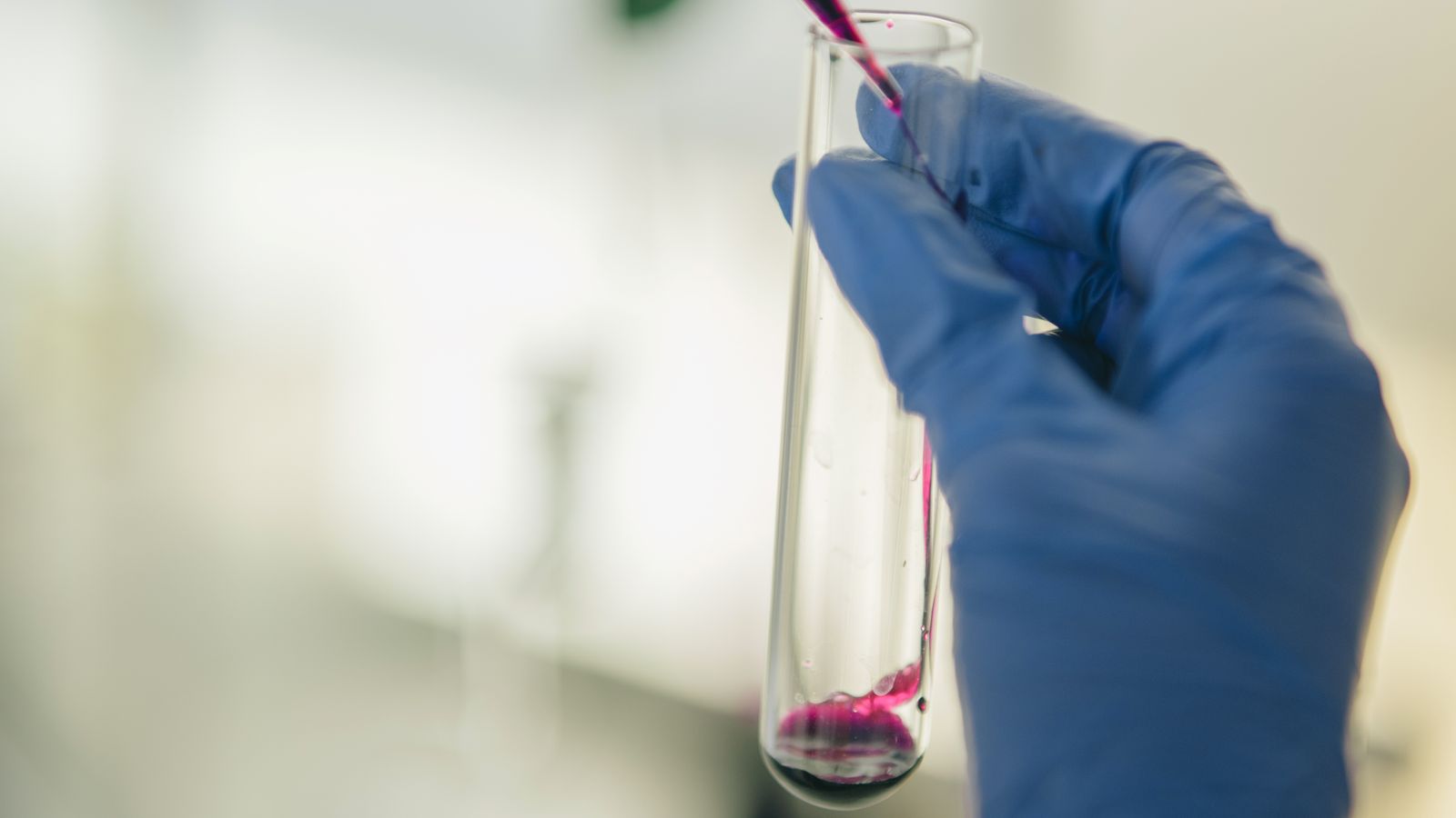 A gloved hand holds a dropper above a test tube on a laboratory bench.