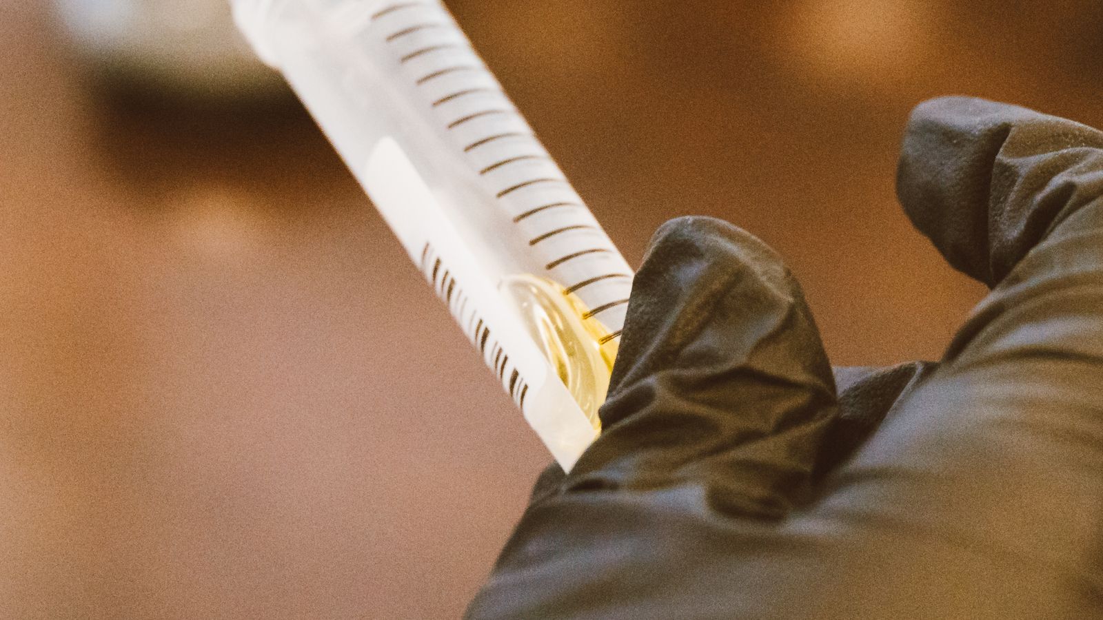 A gloved hand holds a small glass vial up for inspection under laboratory lighting.