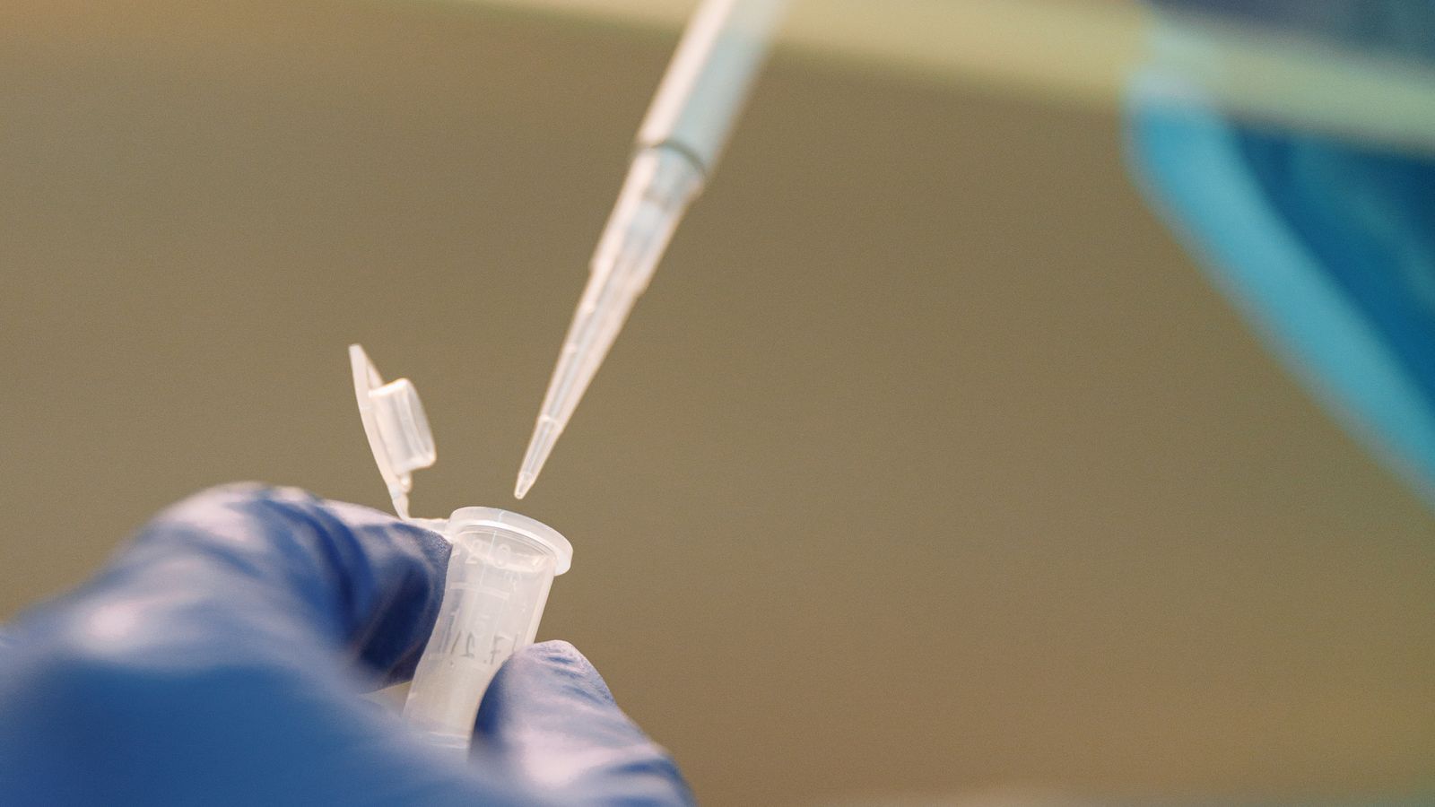 A gloved scientist's hands using a pipette over lab equipment in a modern research setting.