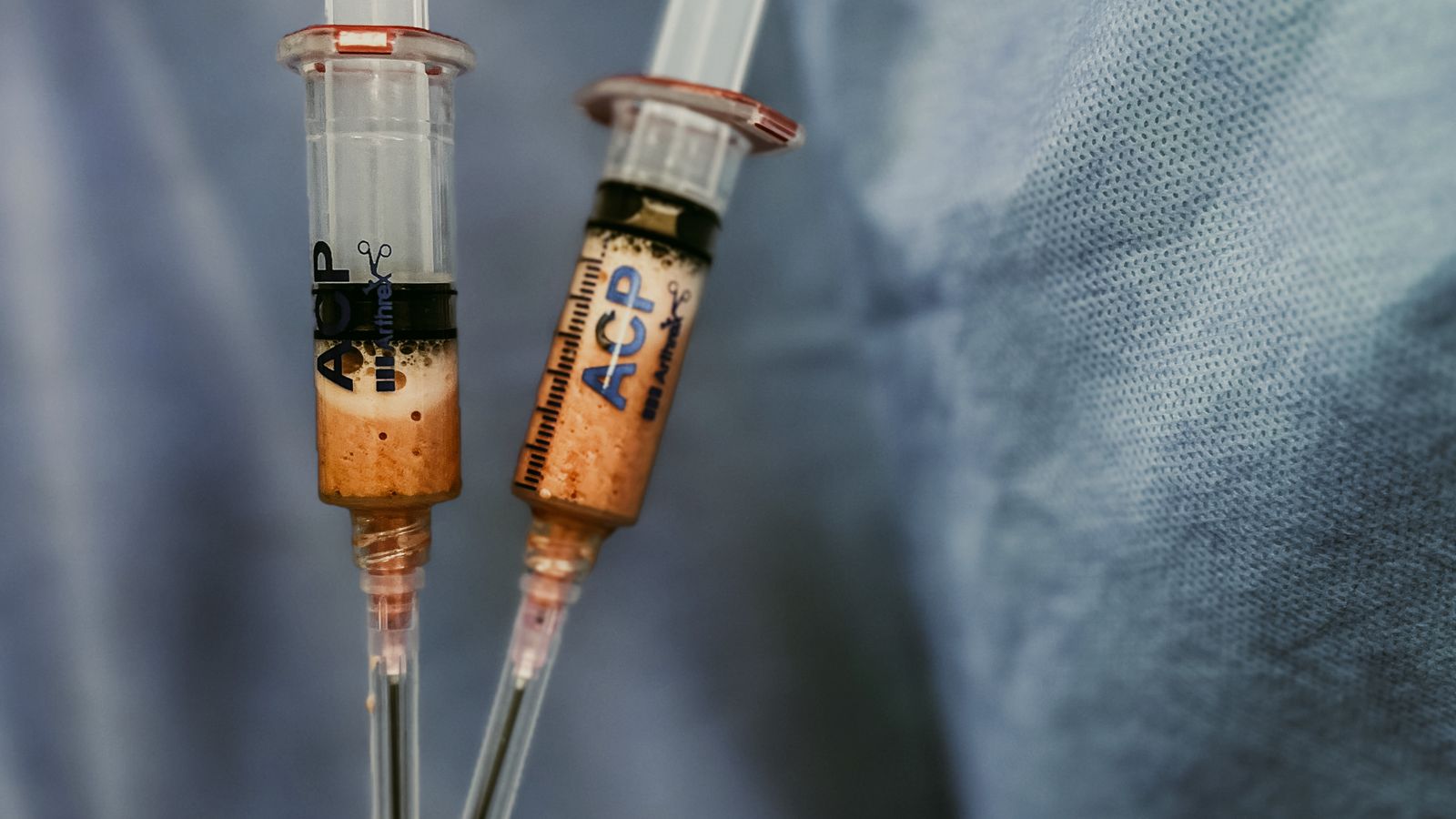 Close-up of gloved hands holding several syringes filled with fluid against a blue background.
